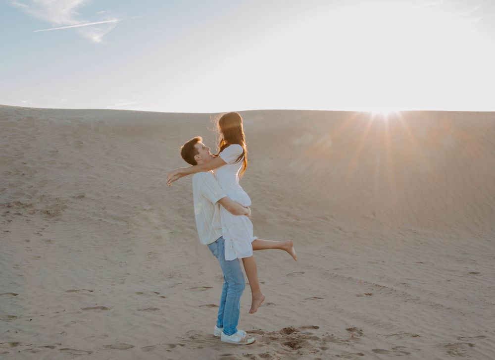 A couple shares a playful lift moment at sunset on sandy dunes.