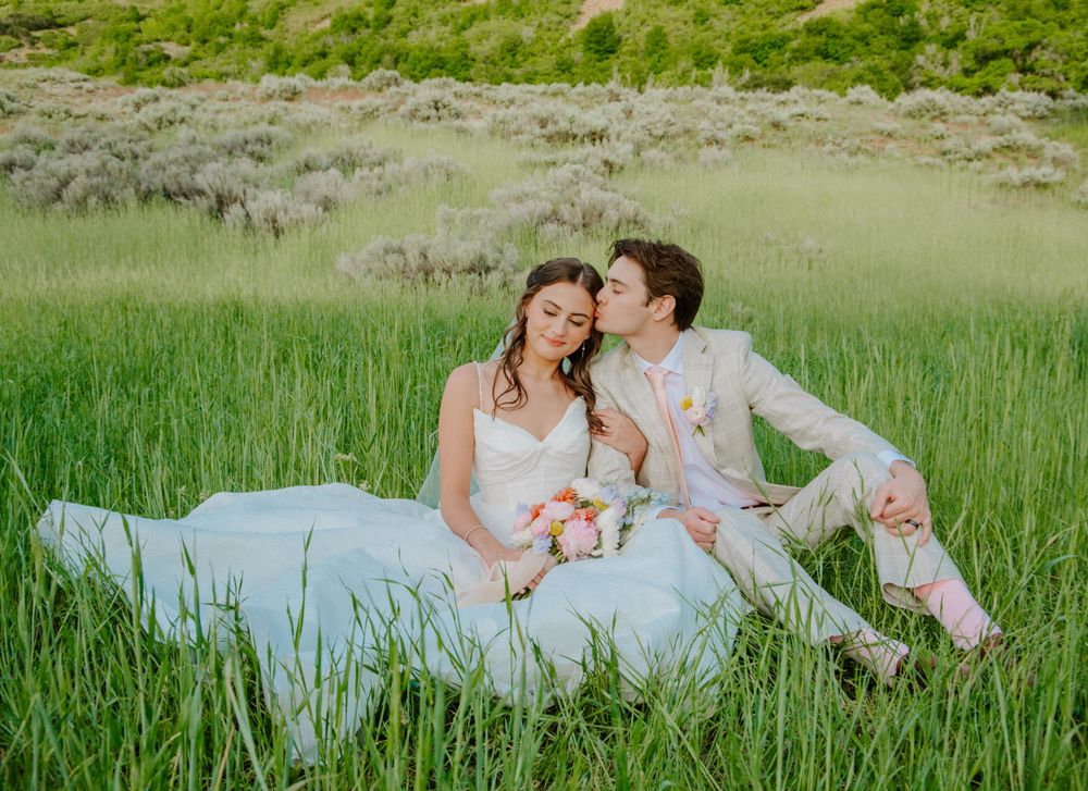 A romantic wedding portrait of a couple sitting in tall green grass with mountain views.