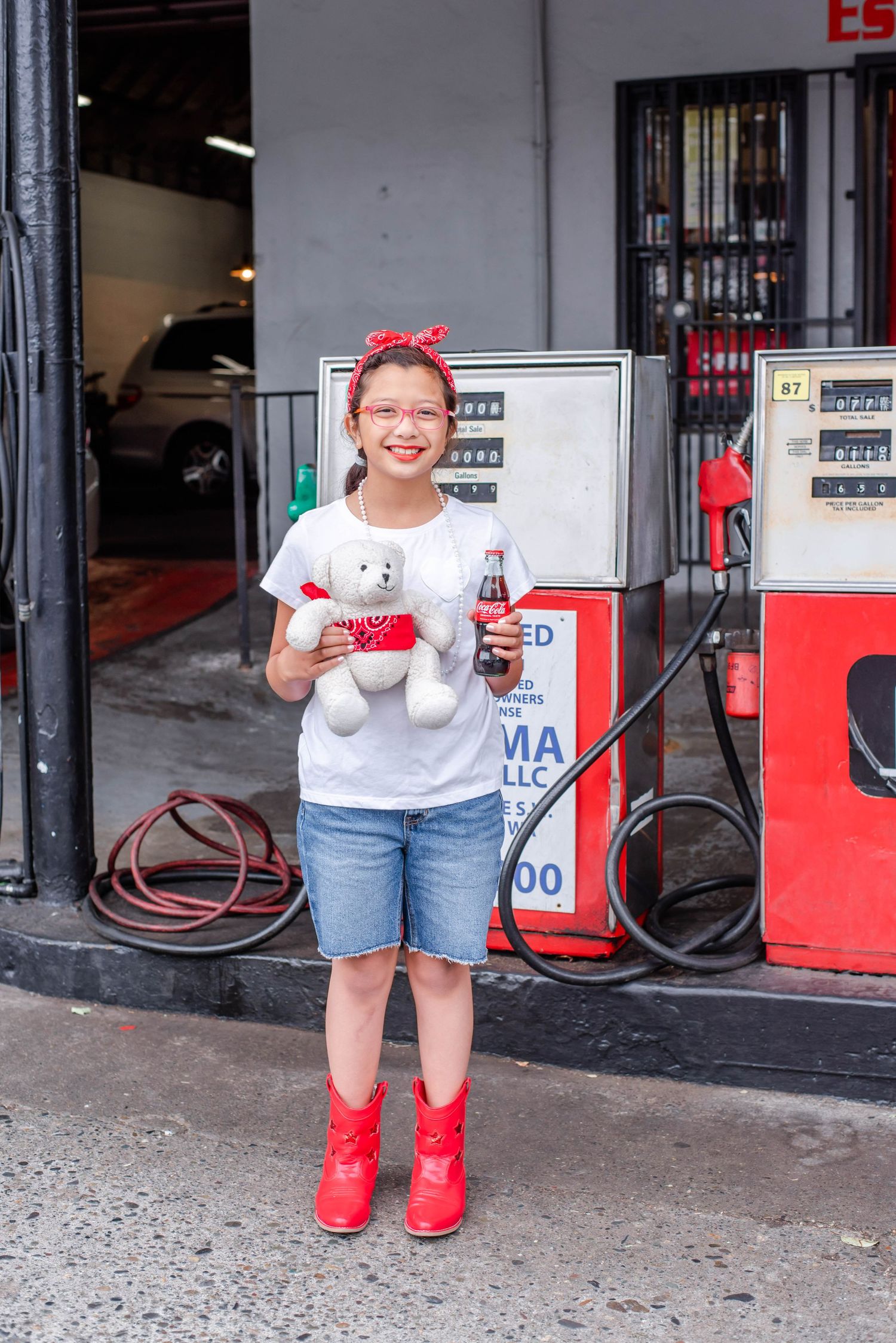 Young child wearing red boots and casual clothes standing in front of gas station pumps.