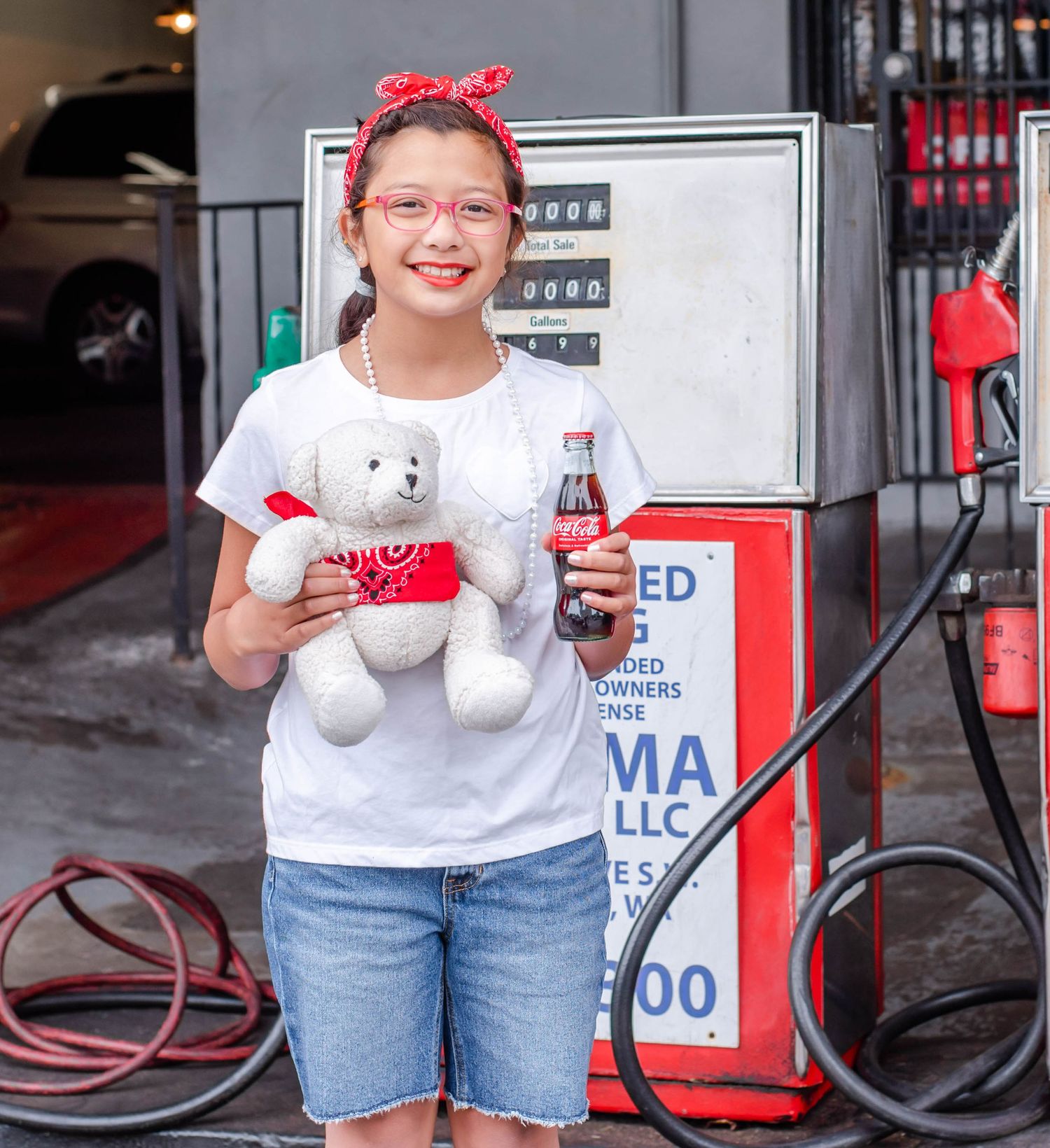 Child in denim shorts and red accessories poses near gas station equipment.