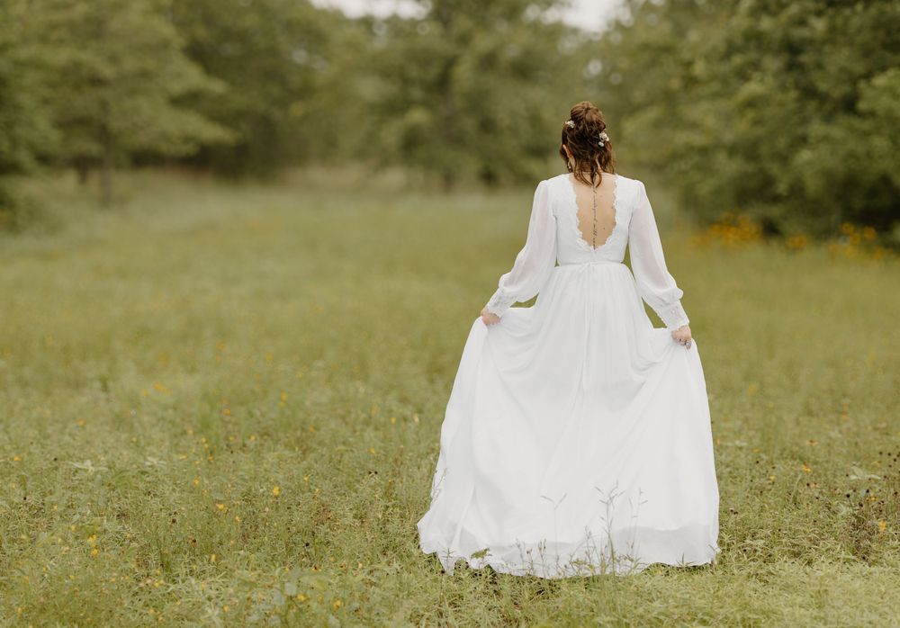 A bride in a flowing white dress stands in a sunlit meadow showing the back of her gown.