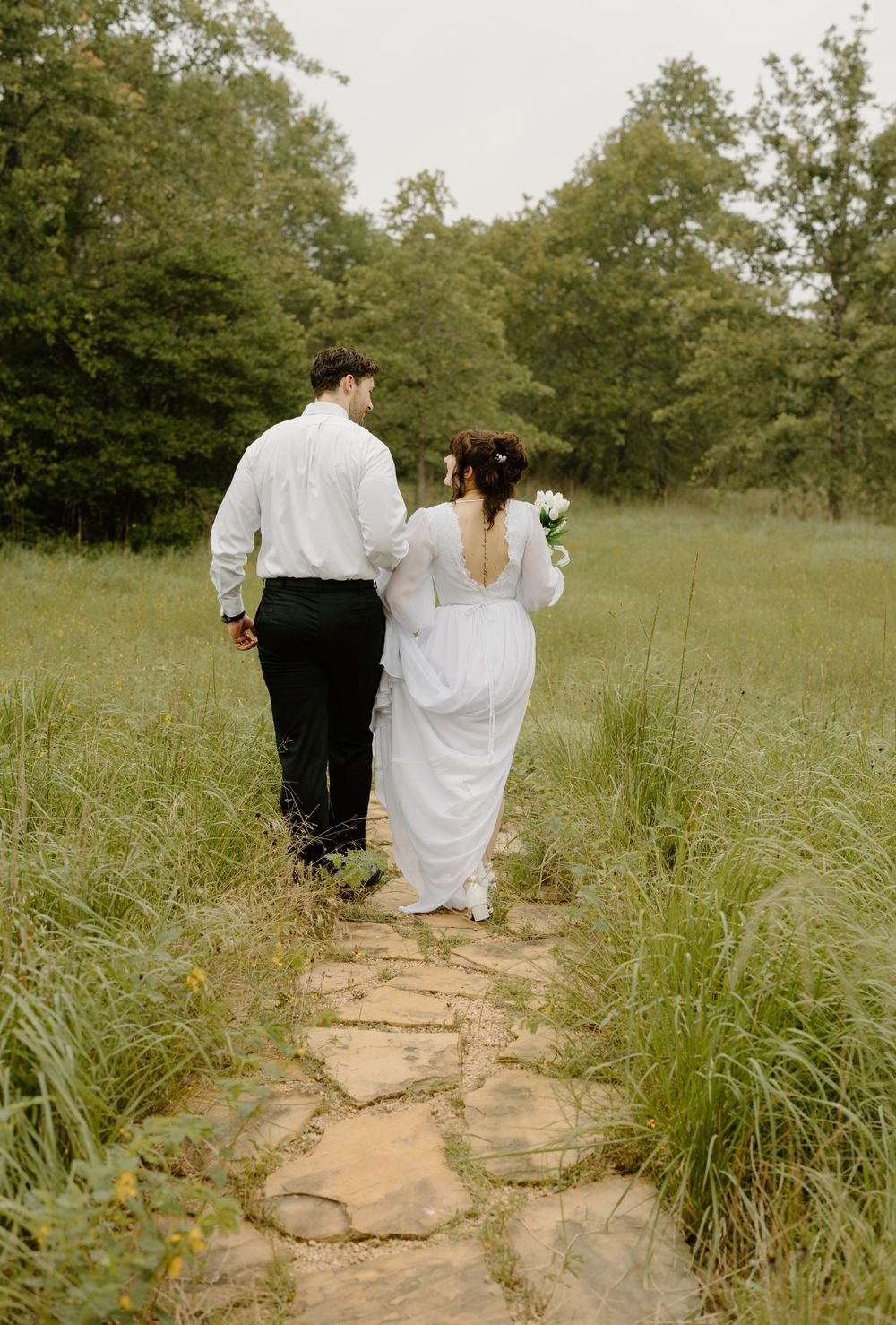 A couple walks together down a stone path through tall grass and trees.