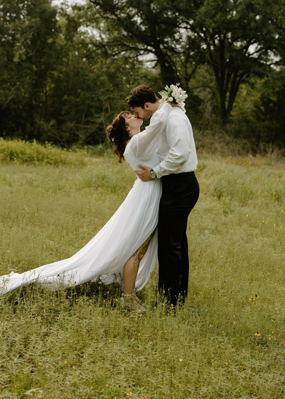 A couple shares an intimate moment in a sunlit field wearing wedding attire.