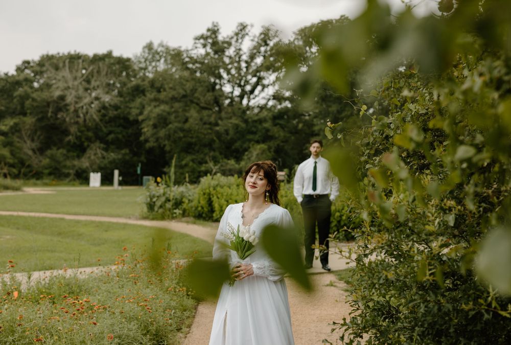 A bride in a garden path while groom walks in the background.