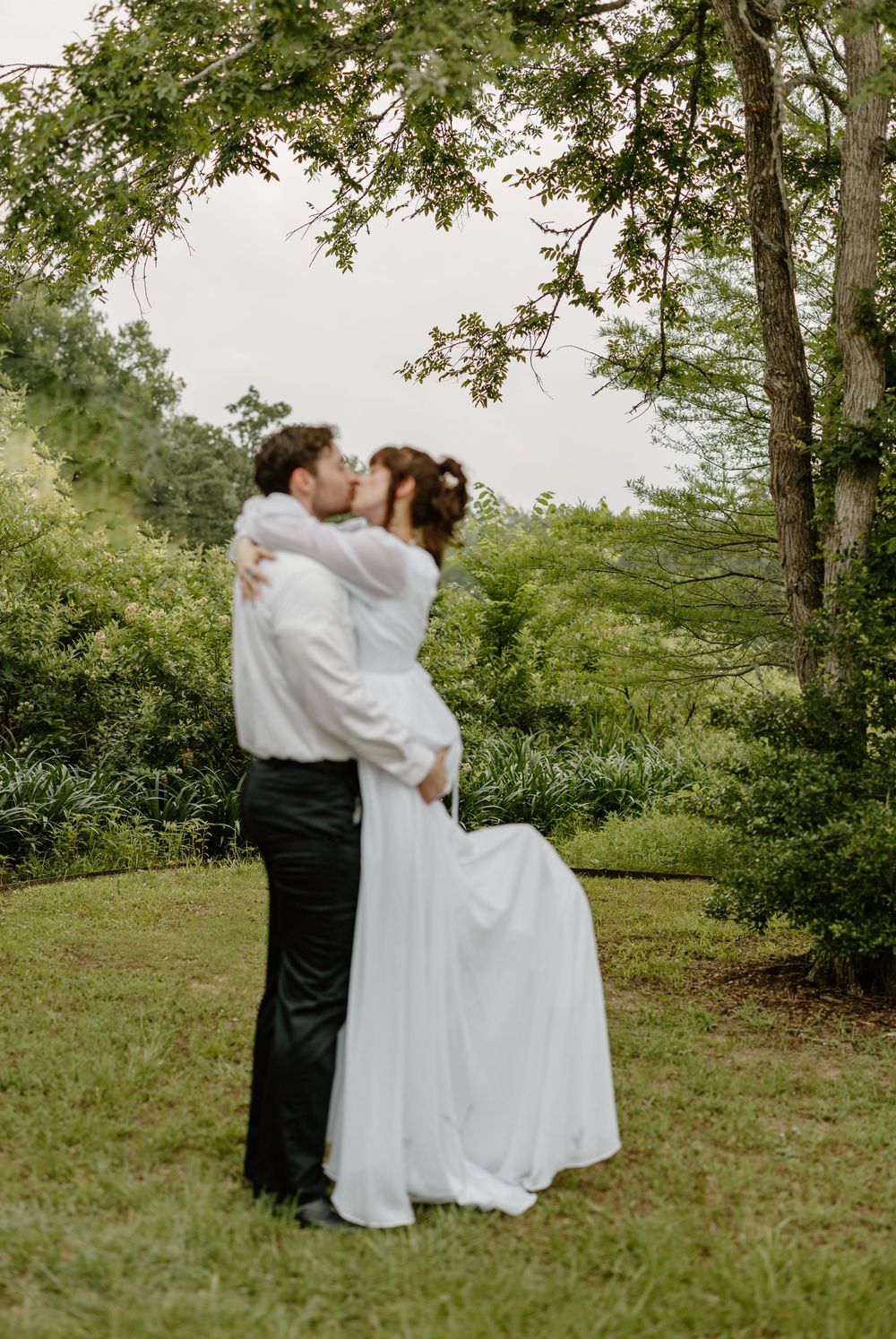 A romantic embrace between newlyweds under a canopy of green trees in a garden setting.
