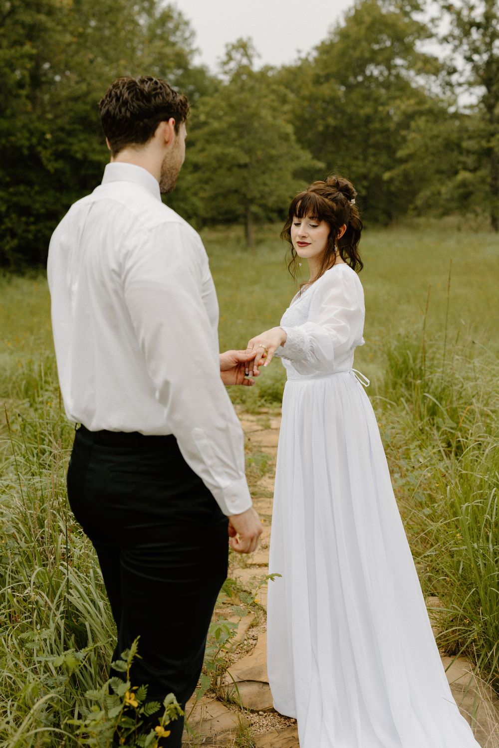 A couple in wedding attire stands on a stone path in a grassy meadow surrounded by trees.
