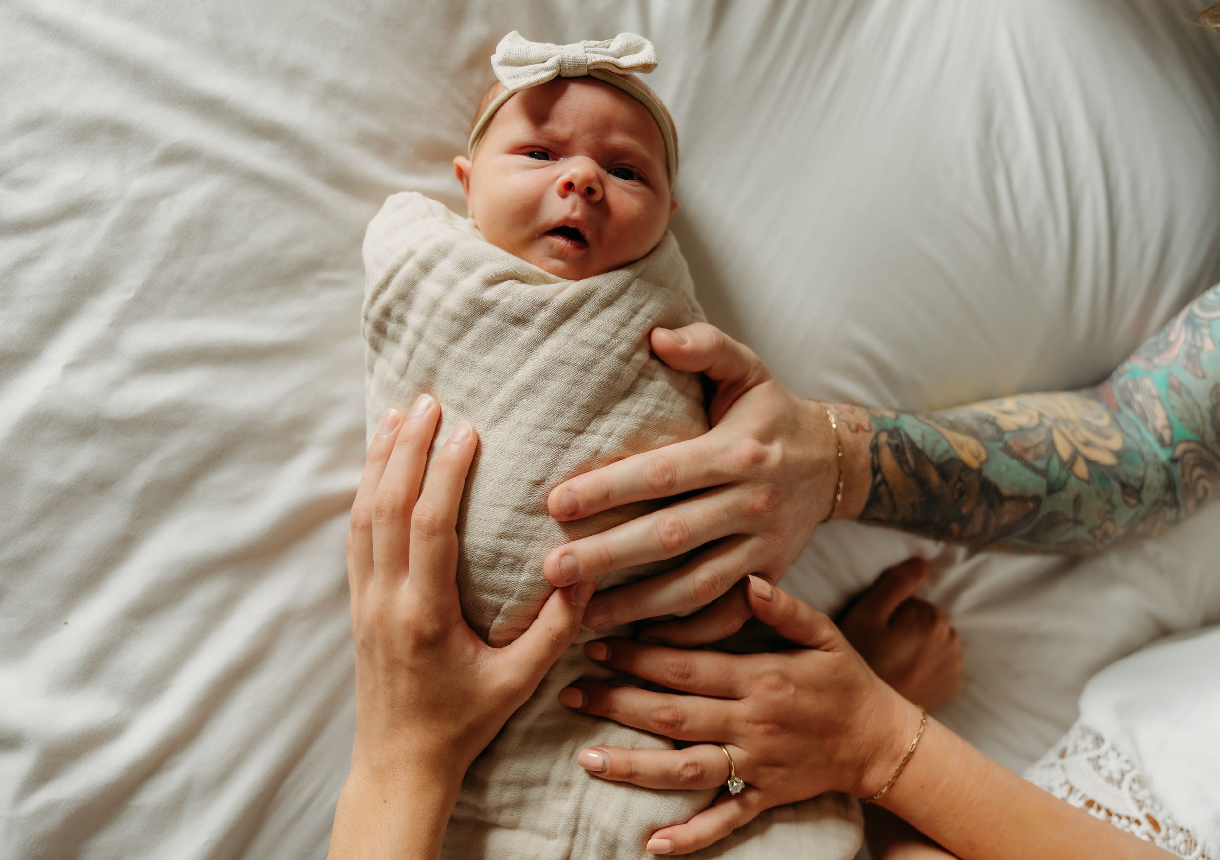Newborn baby in white swaddle wrap with multiple hands reaching to touch.