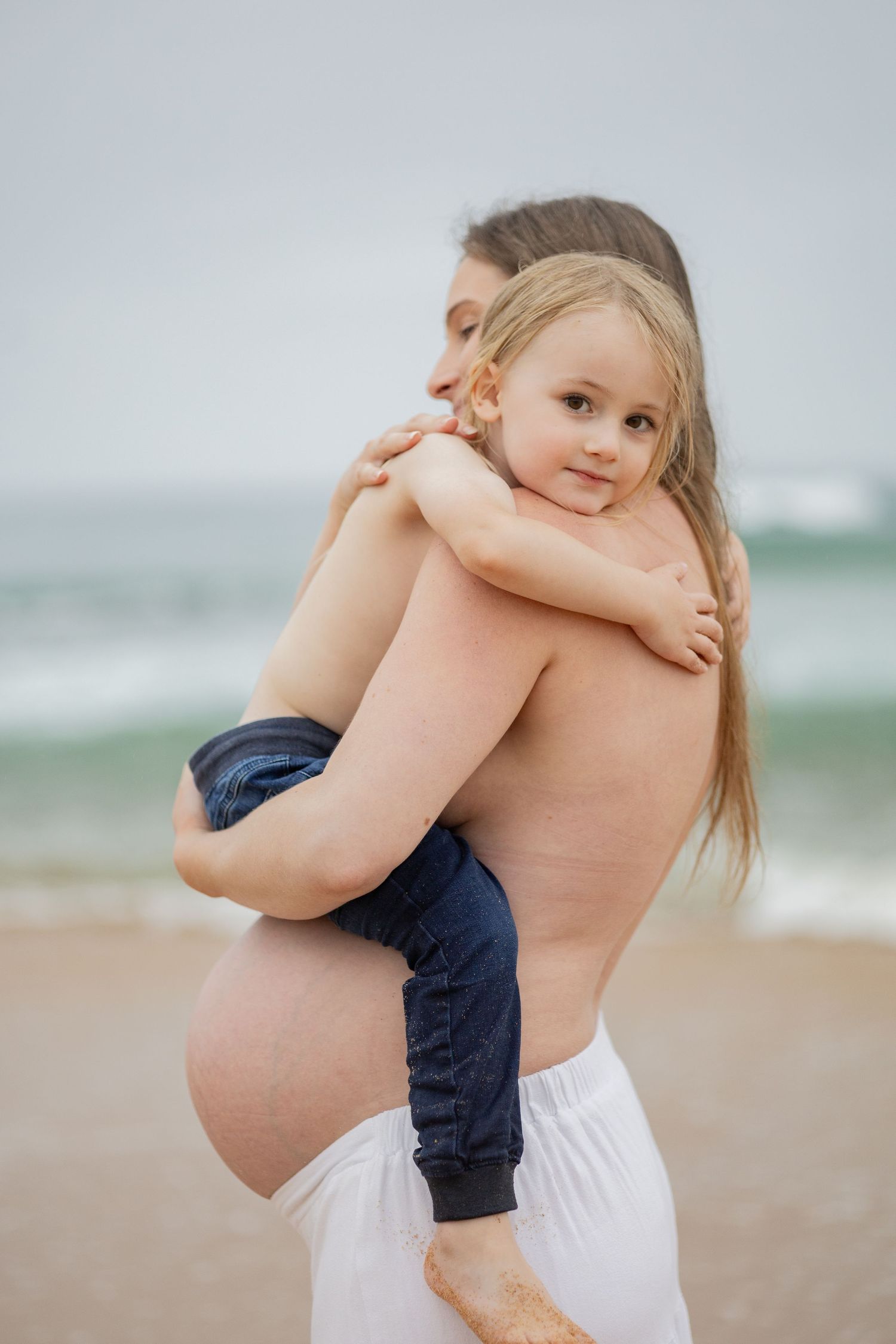 Pregnant mother holding young child on beach with ocean in background.