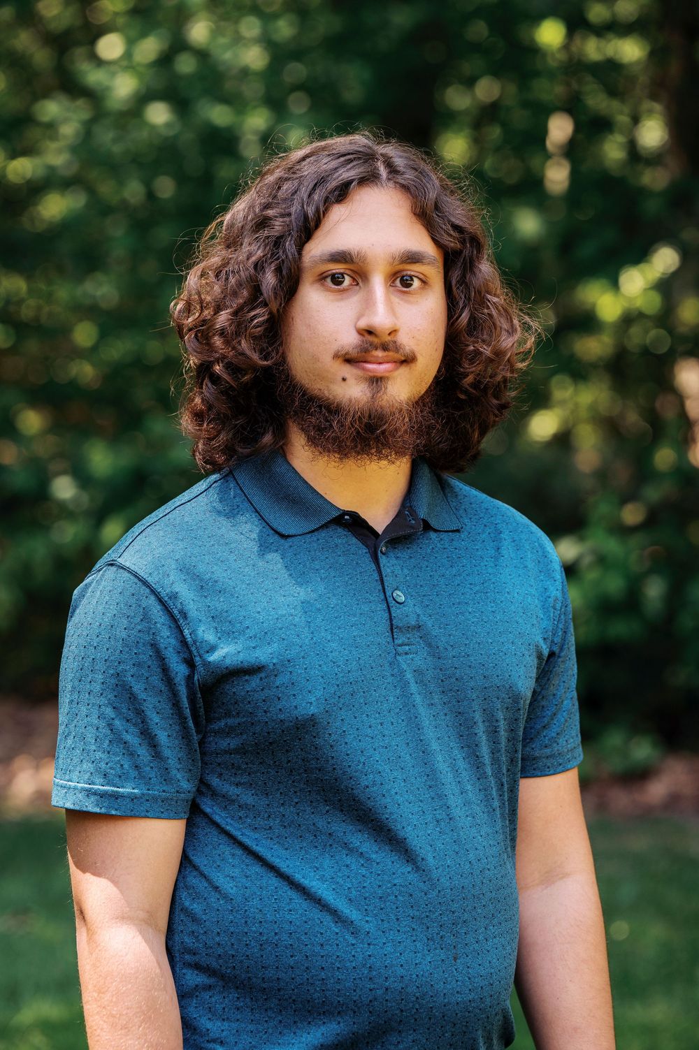 A young man with long curly hair and a beard wearing a teal polo shirt stands in a green outdoor setting.