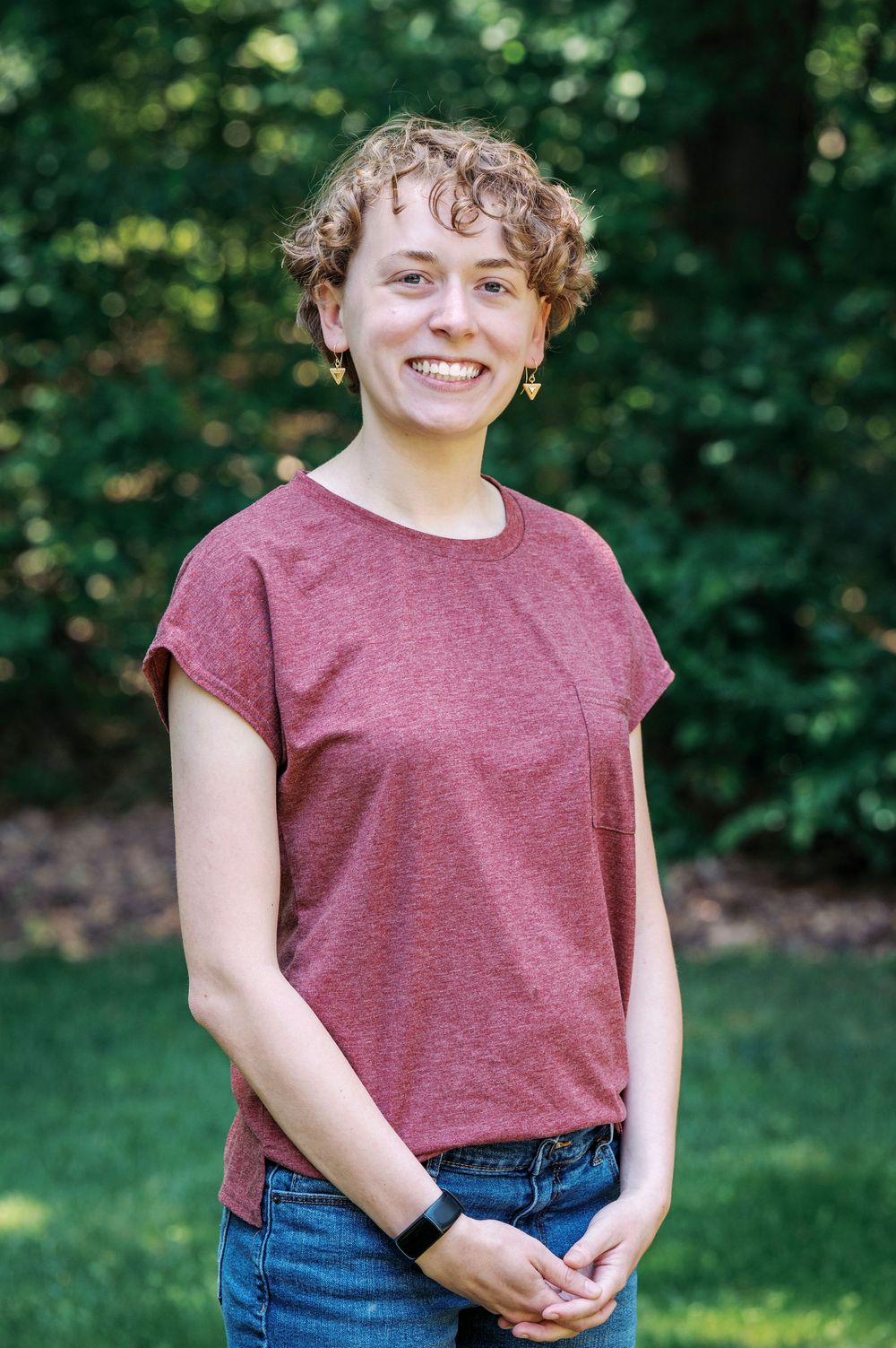 A smiling young person with curly hair wearing a mauve t-shirt and jeans stands in a lush green outdoor setting.