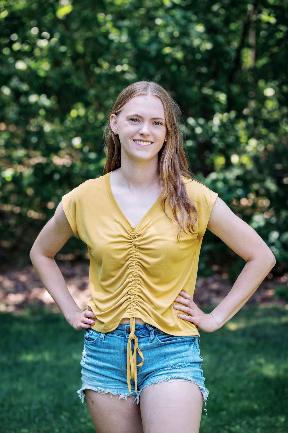 A smiling young woman in a yellow top and denim shorts poses with hands on hips in a sunny outdoor setting.