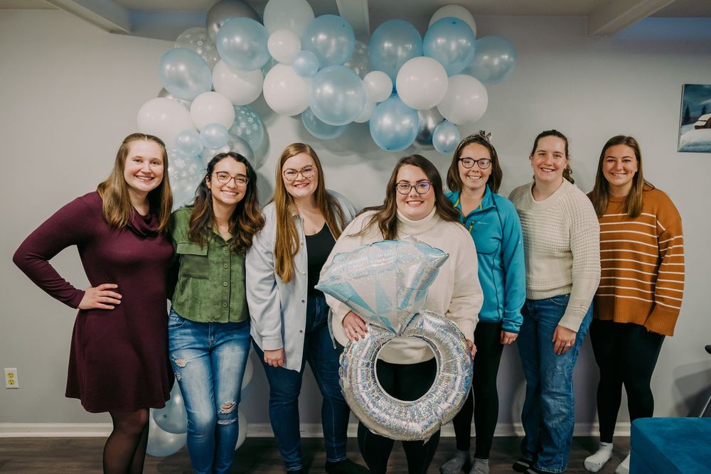 Group of seven women celebrate at bridal shower with blue and white balloon arch and ring balloon.