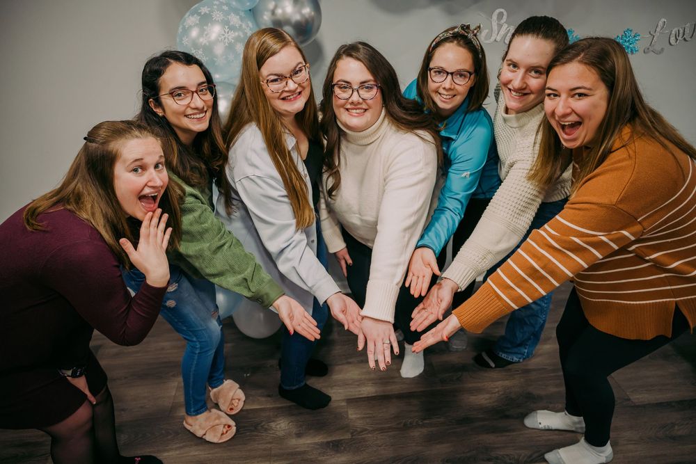 Seven women lean in smiling showing off engagement ring during fun bridal shower celebration photo.