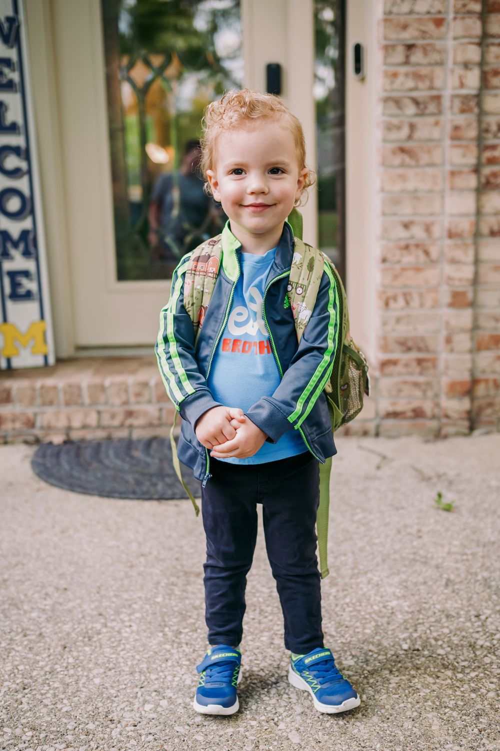 Cheerful redheaded toddler boy poses with backpack outside brick home on first day of school.