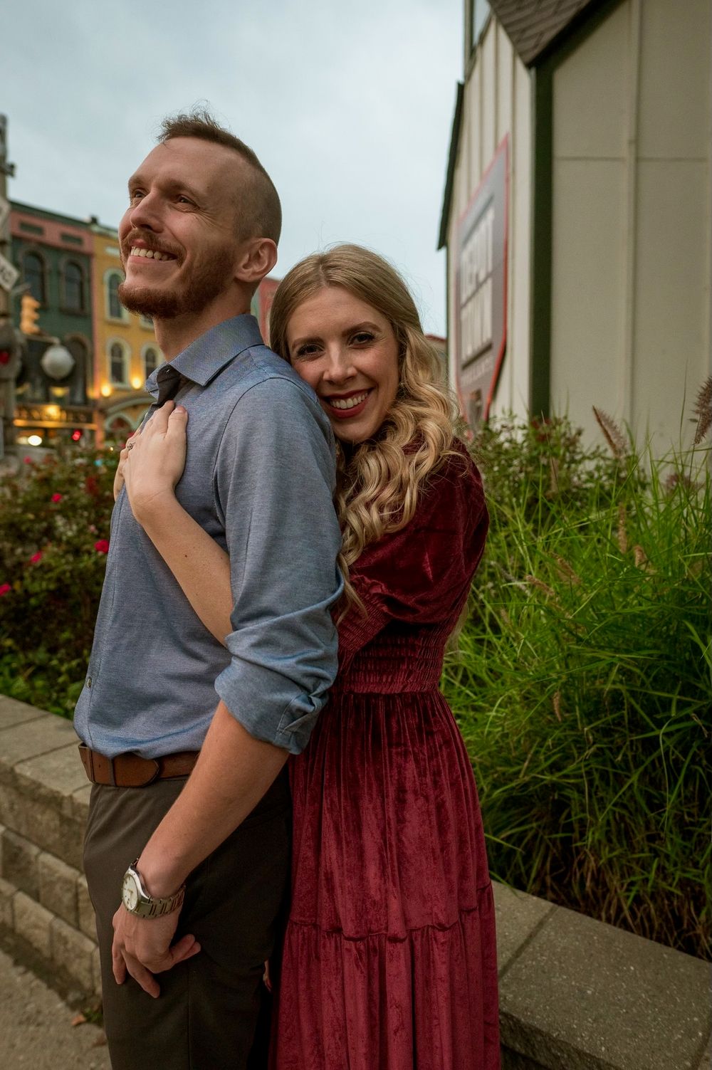 Smiling woman in burgundy velvet dress hugging man from behind on a charming town street.