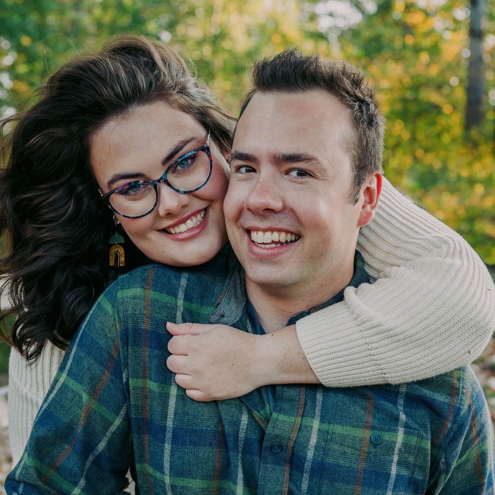 Happy couple smiling outdoors in fall, woman hugging man from behind.