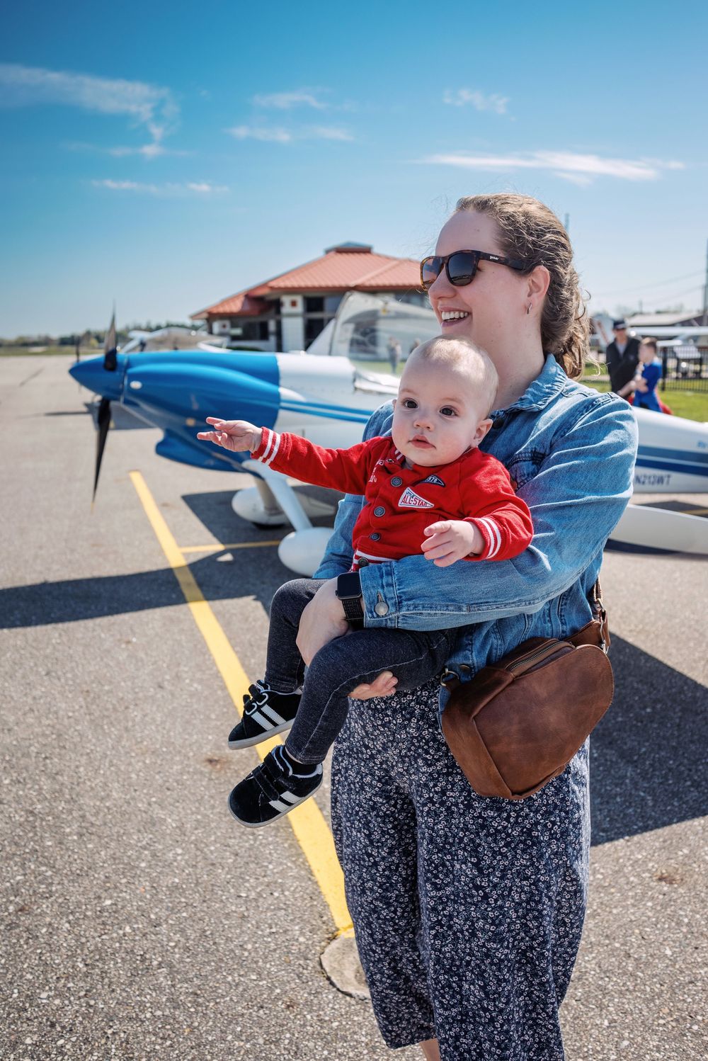 Smiling mother holding baby at airshow with blue plane in background.