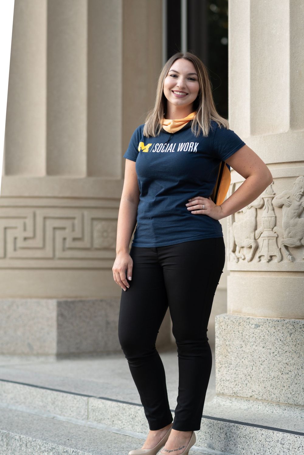 A young woman in a University of Michigan Social Work t-shirt poses confidently beside stone columns on campus.