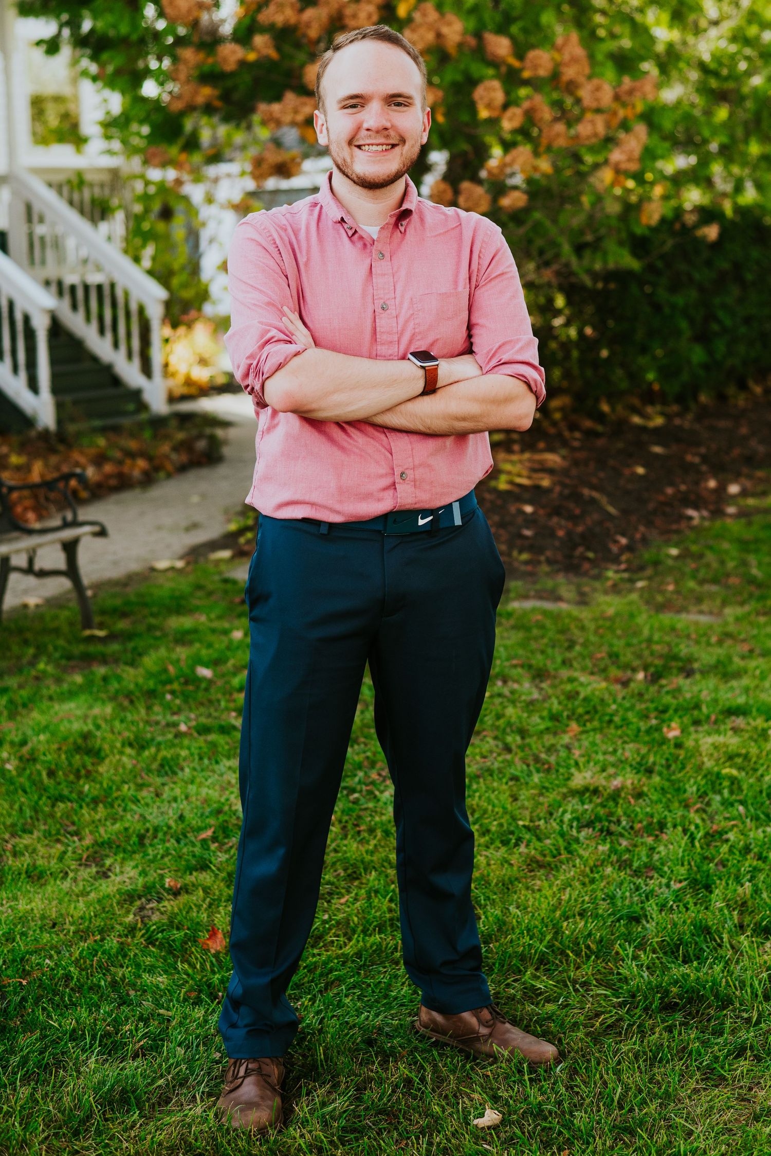 Smiling young man in pink shirt and navy pants posing with arms crossed outdoors.
