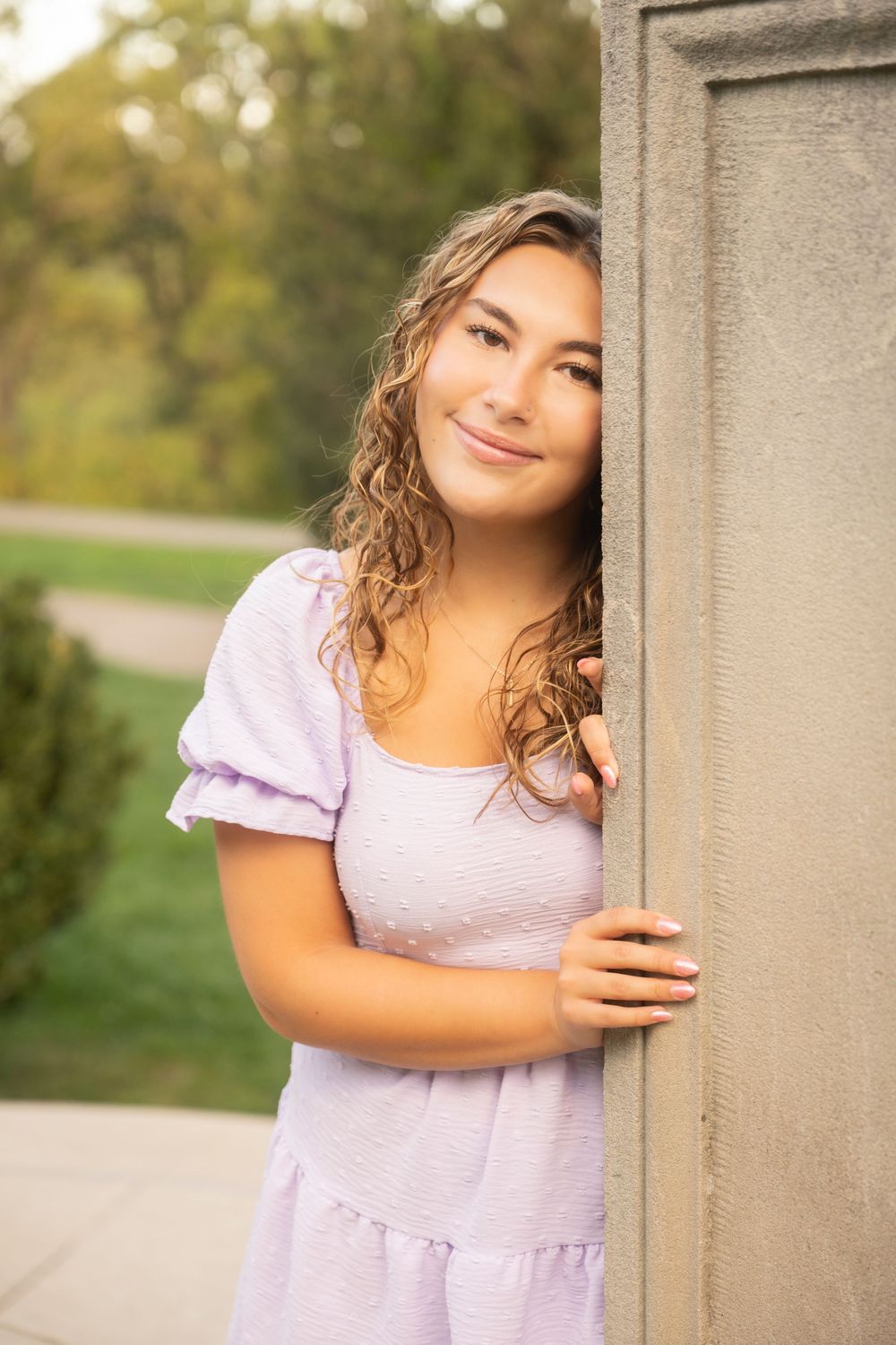 A young woman in a lilac dress with wet hair poses against a stone pillar outdoors in warm sunlight.