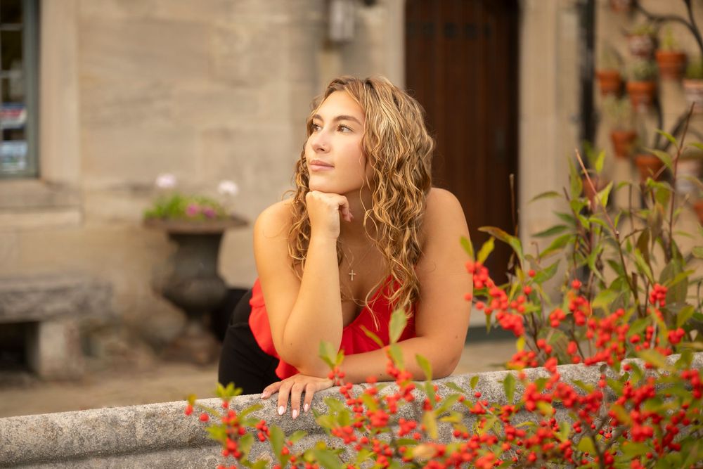 Woman in red dress poses thoughtfully beside vibrant red flowers on stone steps during a photo session.