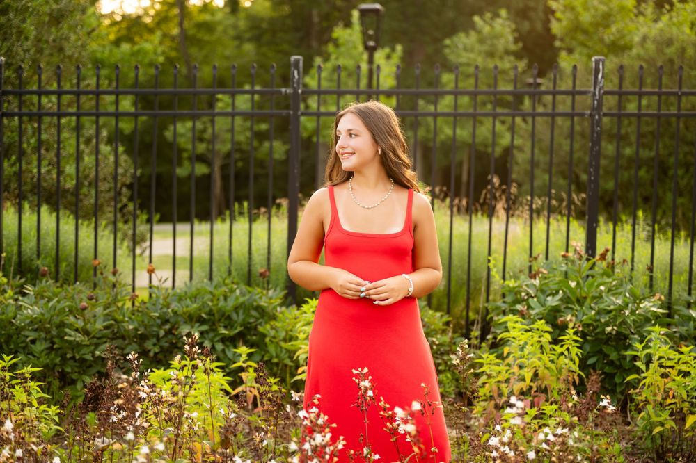 A woman in a vibrant red dress poses gracefully in a lush garden setting with flowering plants and an iron fence.