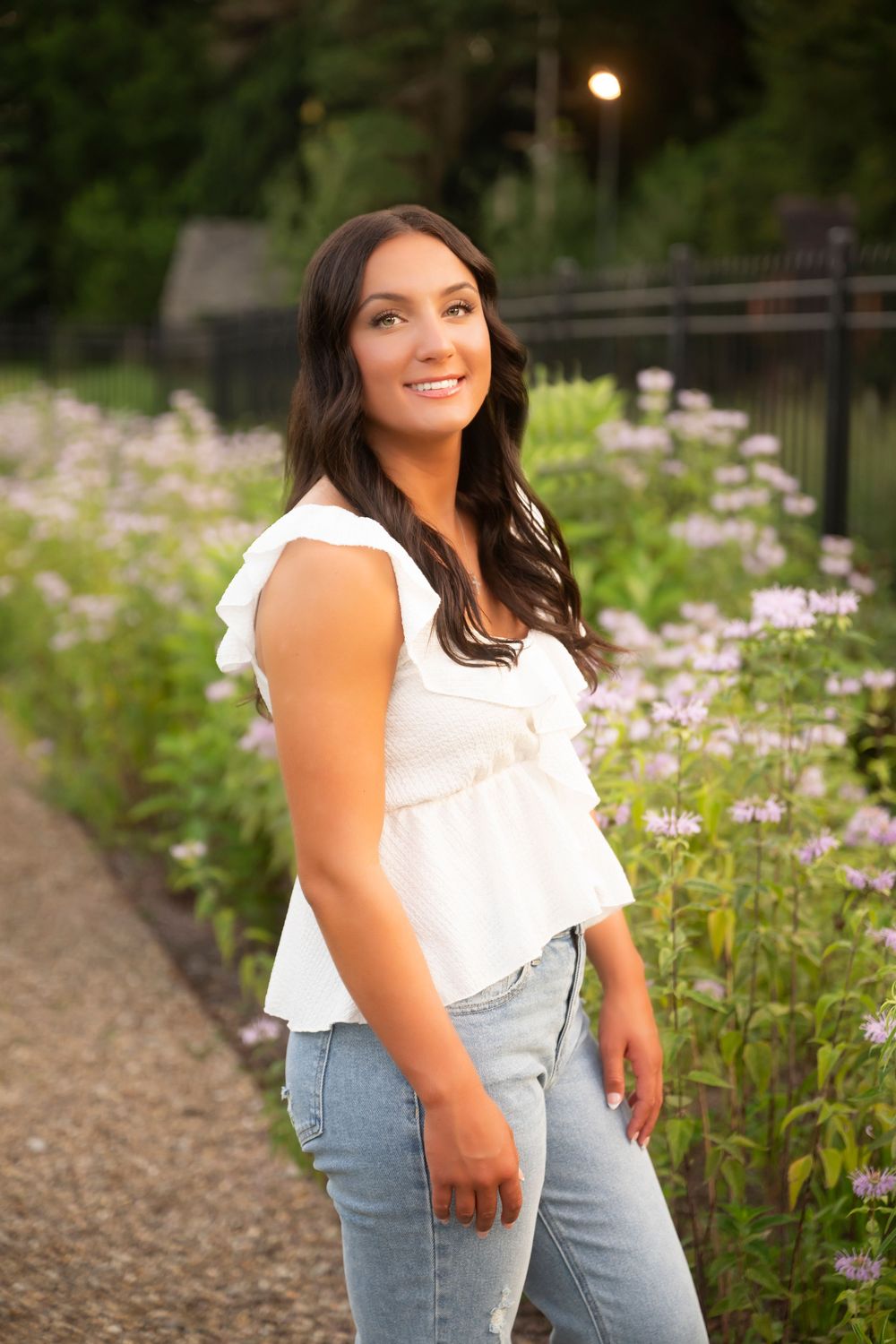 A person in a white sleeveless top and jeans poses along a garden path with blooming purple wildflowers at dusk.