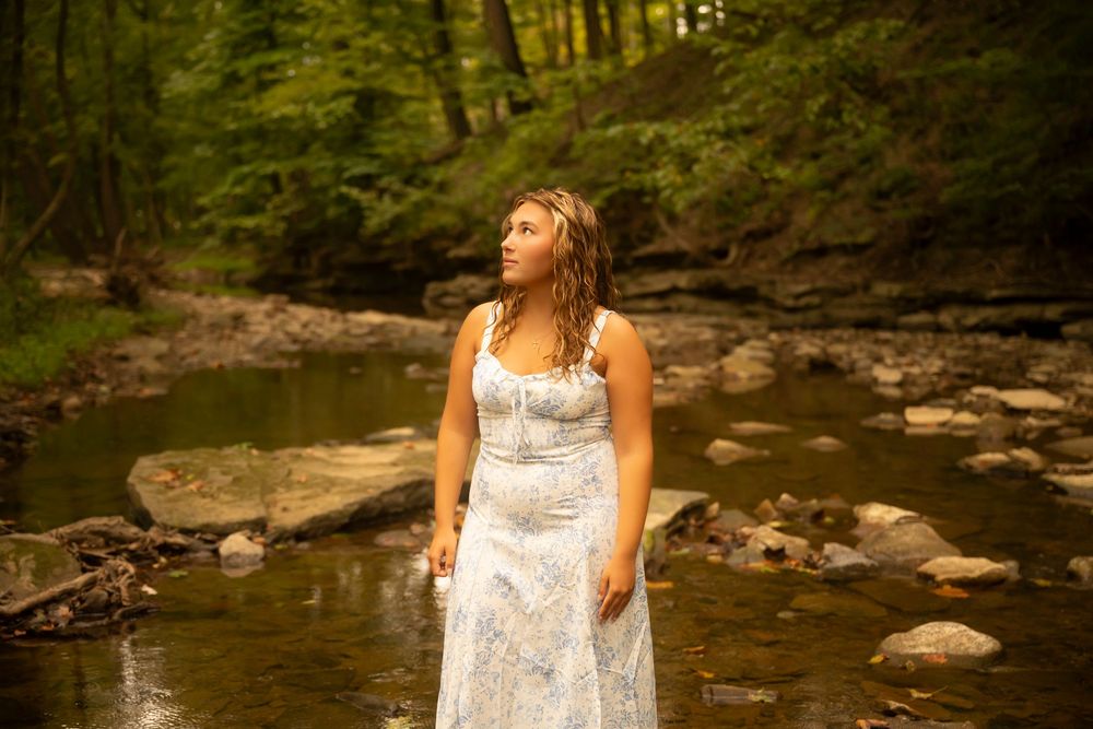 A person in a white dress stands in a shallow stream surrounded by rocks and forest scenery.