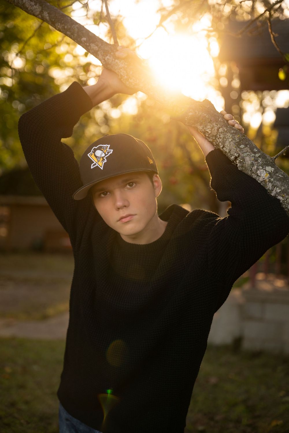 Silhouette of a person in a baseball cap and black hoodie poses against golden sunset light filtering through trees.