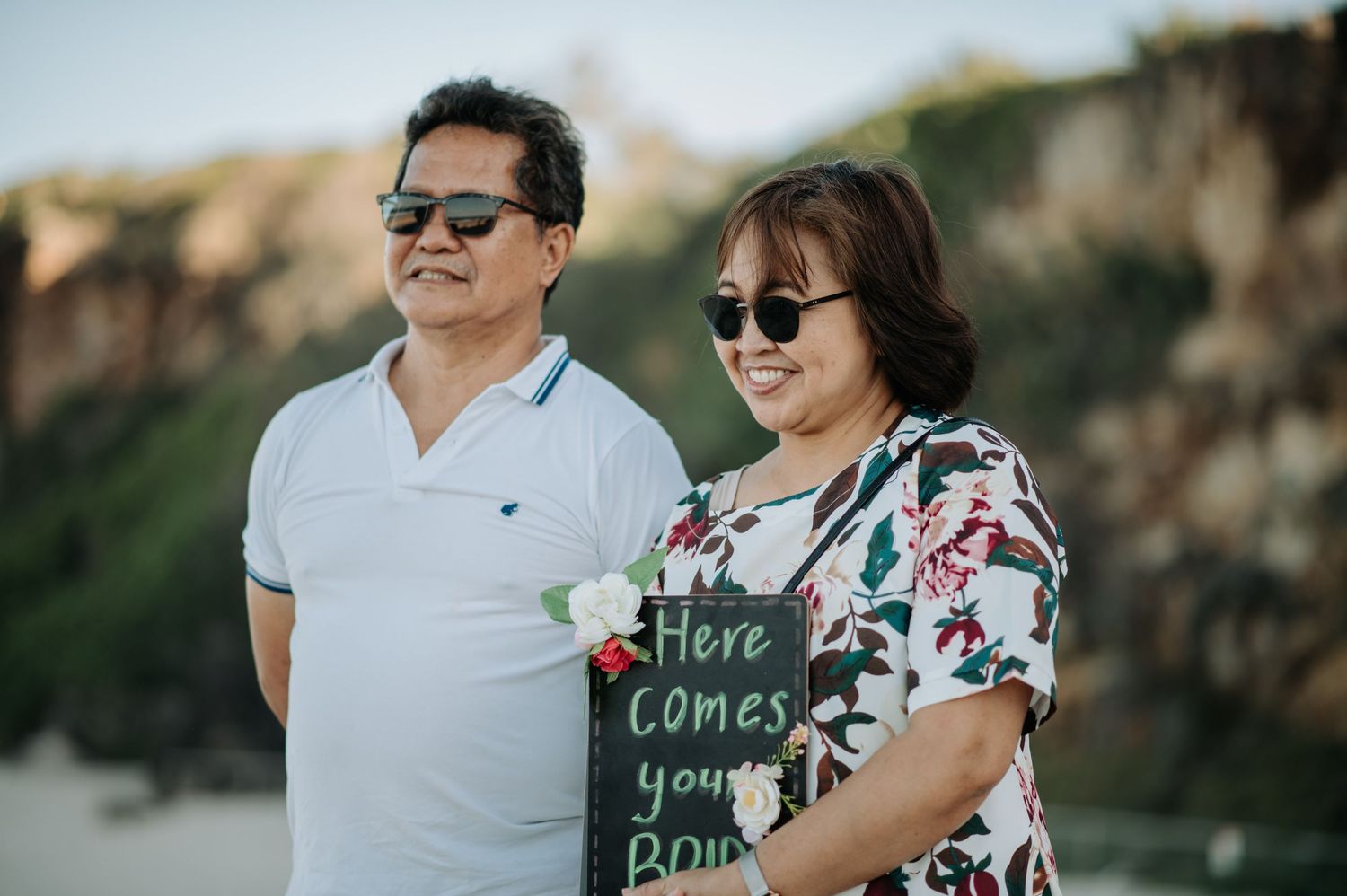 Two people holding a chalkboard sign reading 'Here comes your bride' against a coastal backdrop.