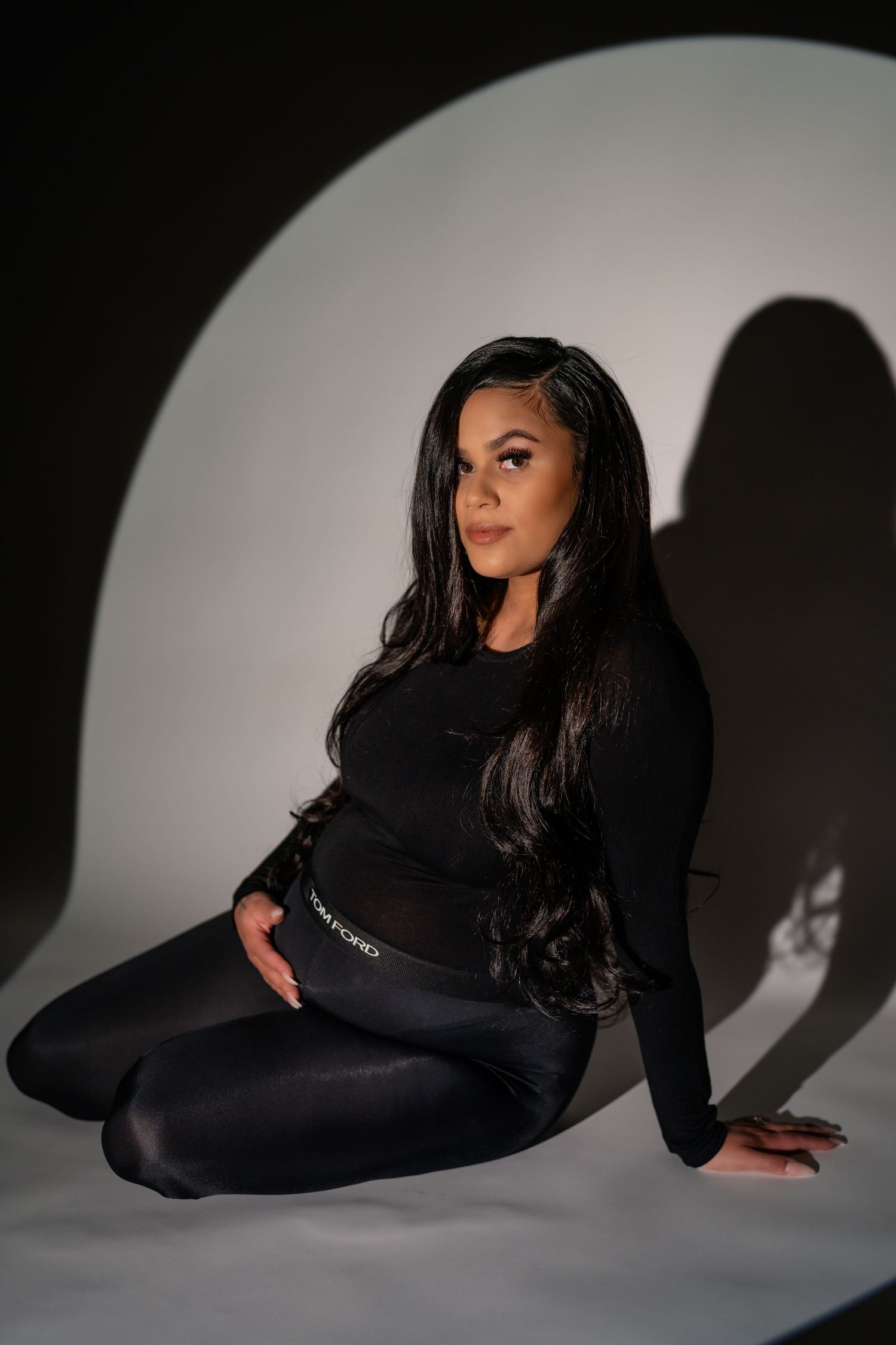 Elegant maternity portraits on light backdrop feature poses in a fitted black dress with dramatic shadows.
