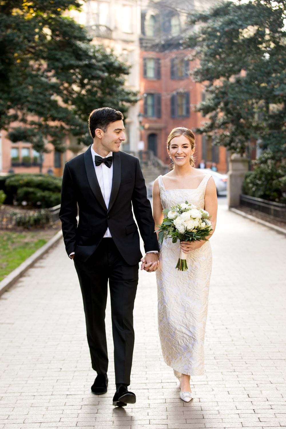Couple strolls hand in hand along brick pathway surrounded by lush greenery.