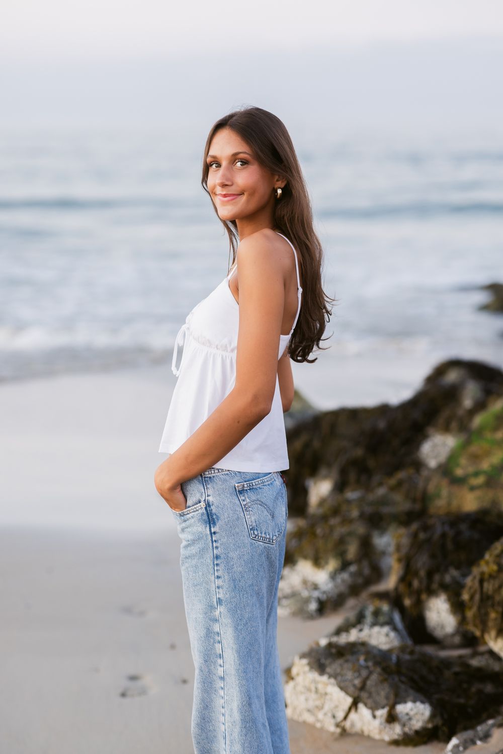 Young lady with brown hair wears white tank top and blue jeans posing sideways near ocean's edge at sunset. Exeter senior photographer
