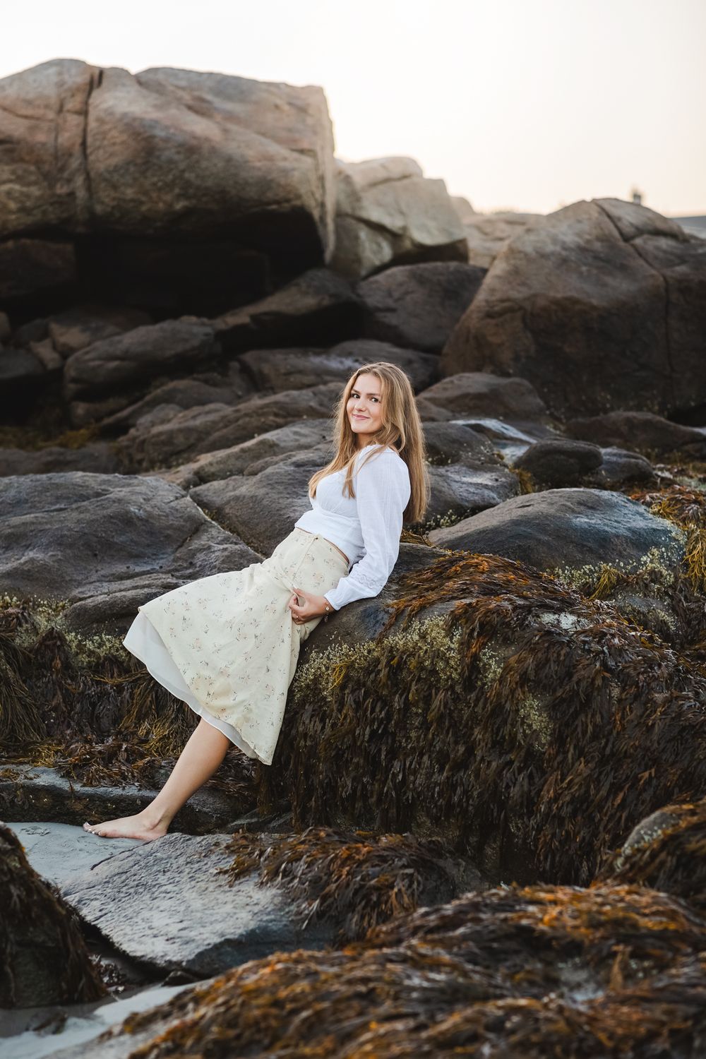 Senior girl wears white blouse and yellow floral skirt leaning on dark rocks at Wingaersheek Beach in Gloucester, North Shore MA.
