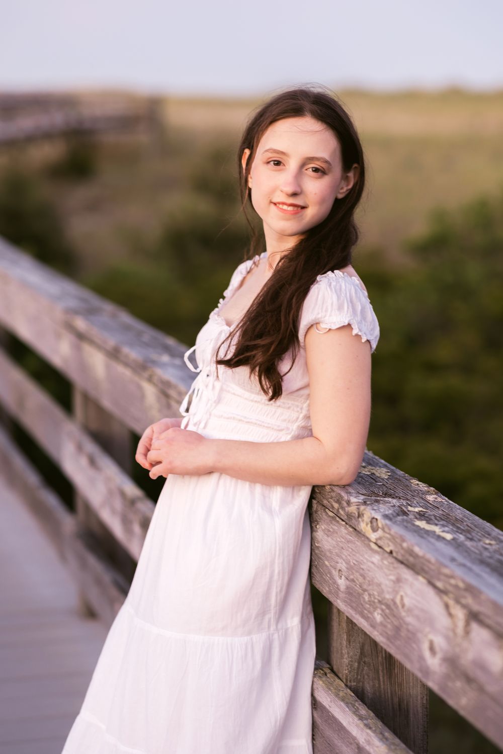 Senior girl in white dress leans on wooden boardwalk railing at blue hour with coastal grass behind her. Seacoast NH senior photographer