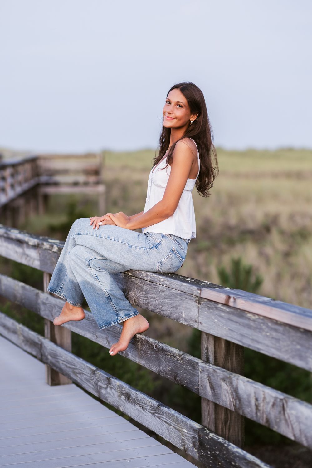 Young lady with brown hair wears a white tank and blue jeans for senior photos sitting on weathered gray boardwalk with coastal seagrass.