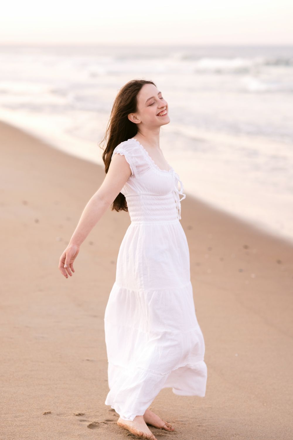 A joyful young woman in a white flowing dress poses barefoot with head back on a sandy beach at sunset. Salisbury senior photos.
