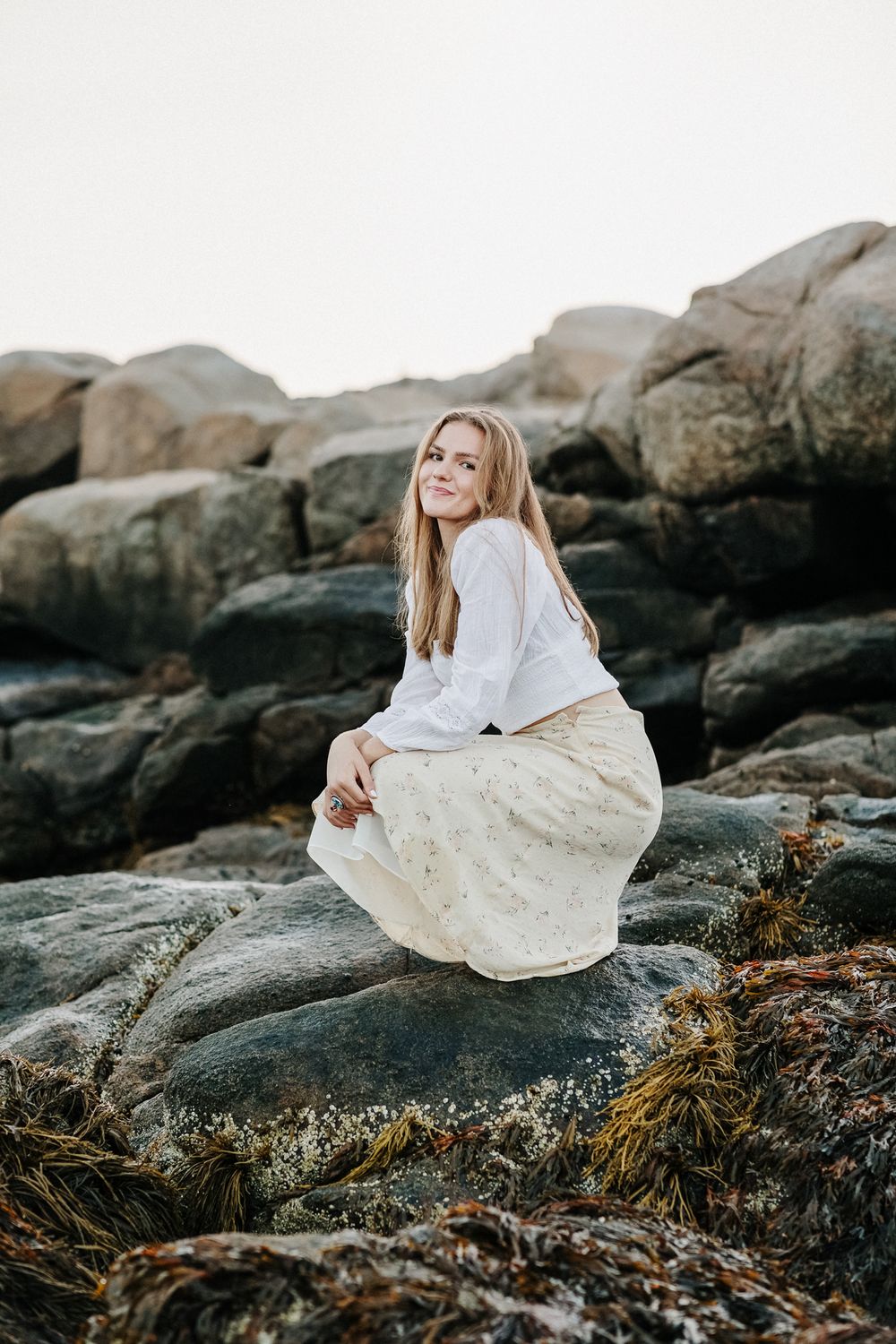 Long hair blond girl wears white blouse and yellow floral skirt crouches on dark rocks at Wingaersheek Beach North Shore for senior photos.