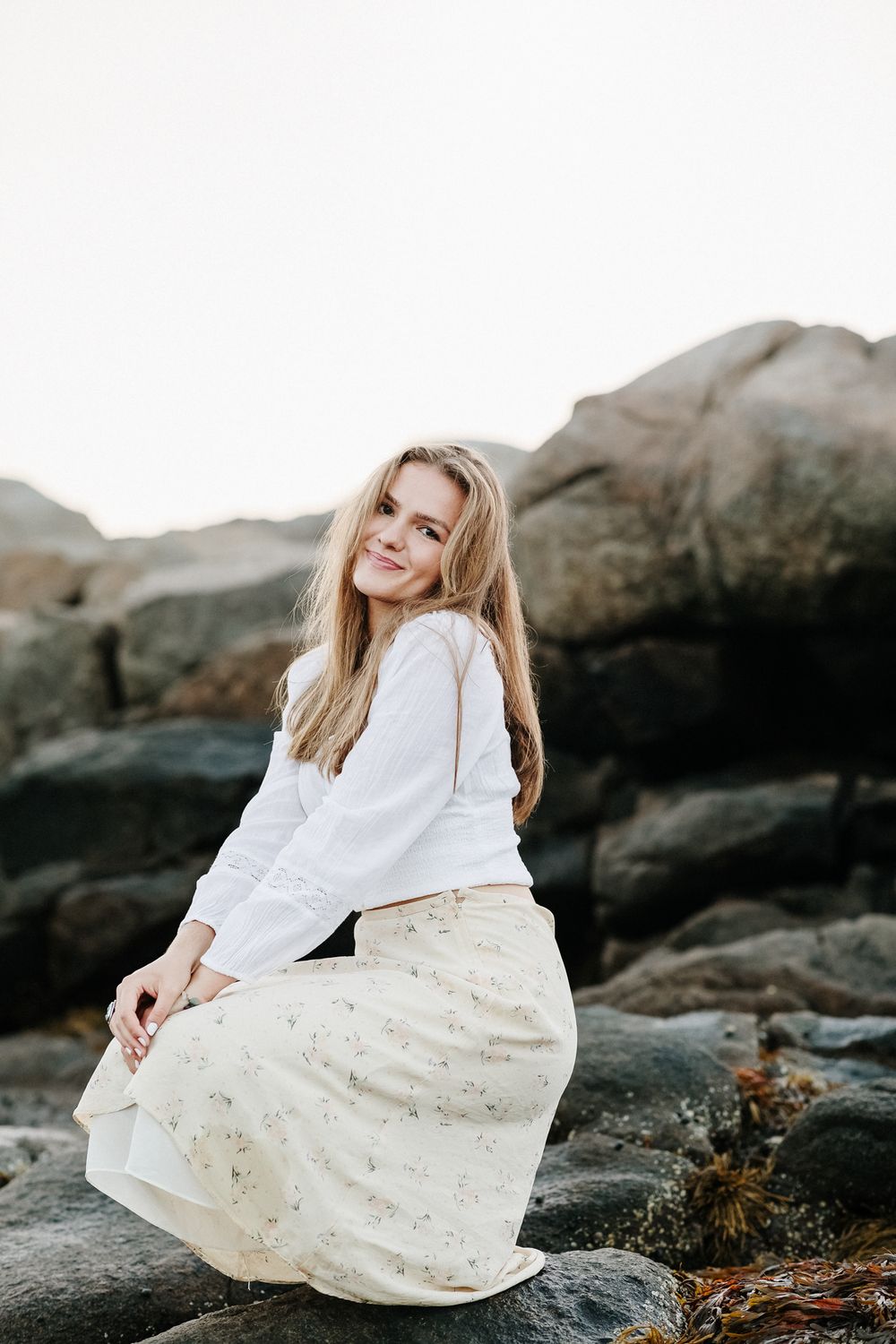 Blond senior girl poses squatting on dark coastal rocks at Wingaersheek Beach in Gloucester North Shore for senior photos.