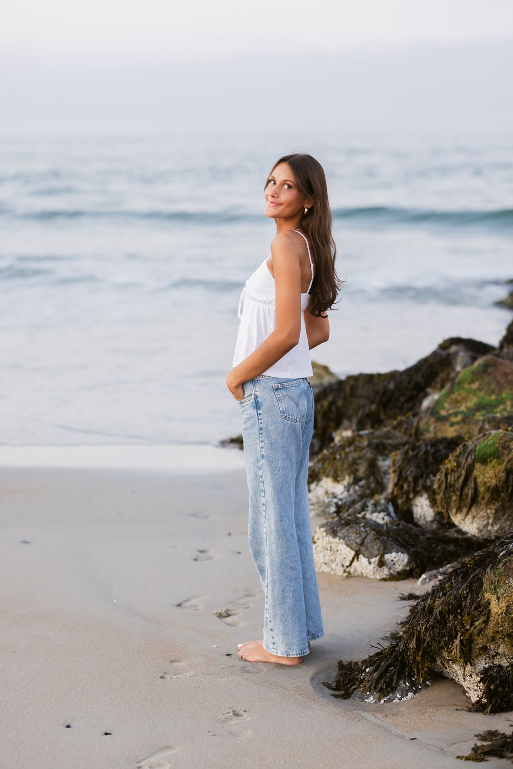Senior girl wears white tank and blue jeans standing sideways by oceans's edge at sunset. Essex North Shore York senior photographer