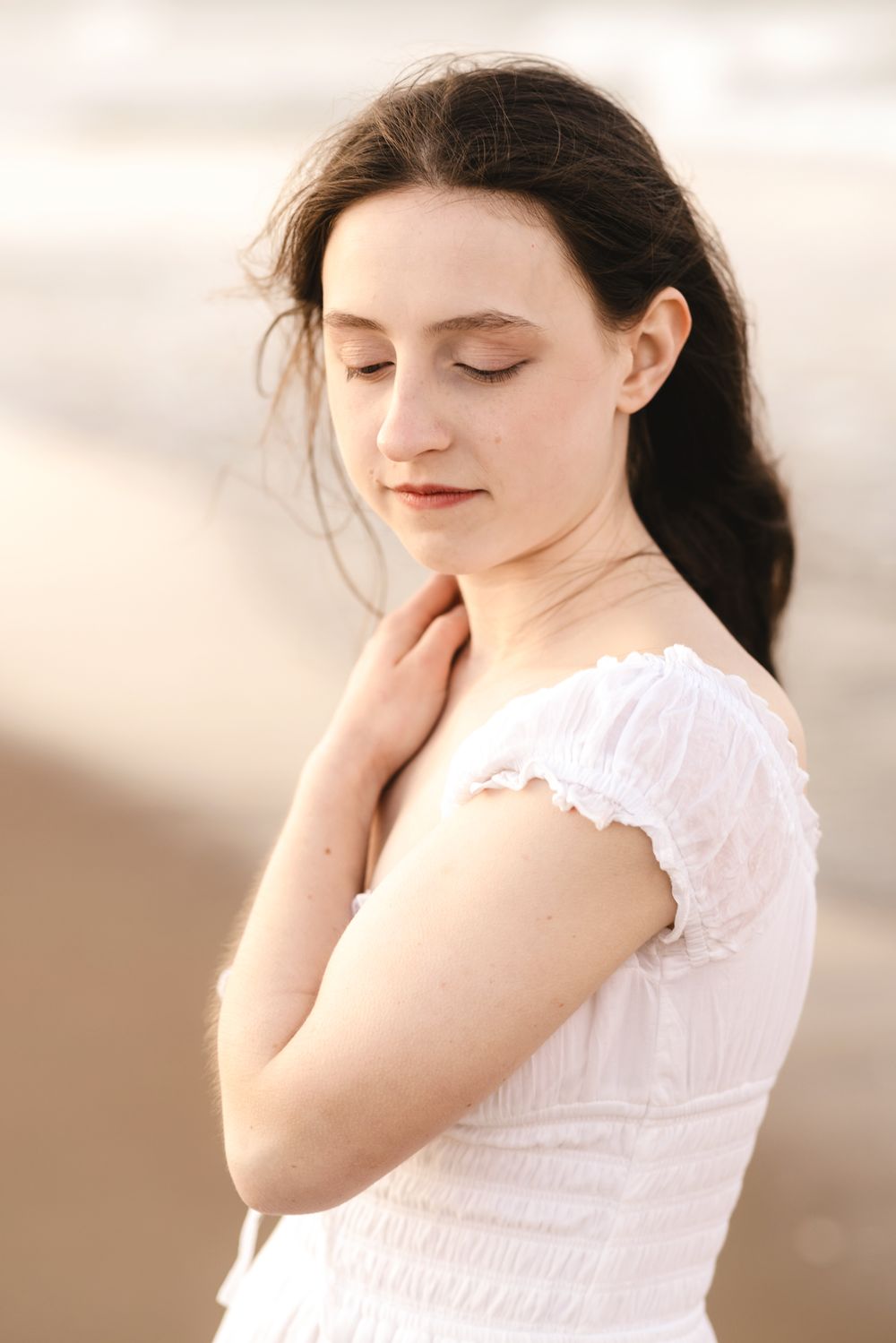 Senior girl with dark hair in white dress, eyes closed, hand on neck, poses at beach for senior photos. Southern Maine senior photographer.