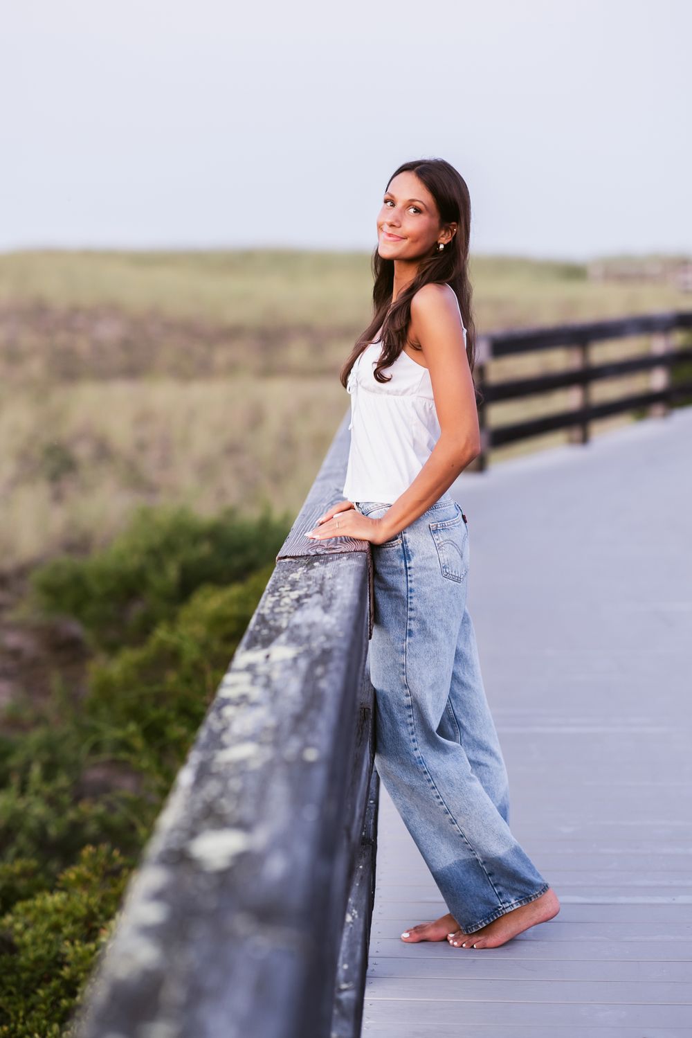 Senior girl wears white tank and blue jeans sits crouching on a weathered boardwalk posing for senior photos. Bedford senior photographer