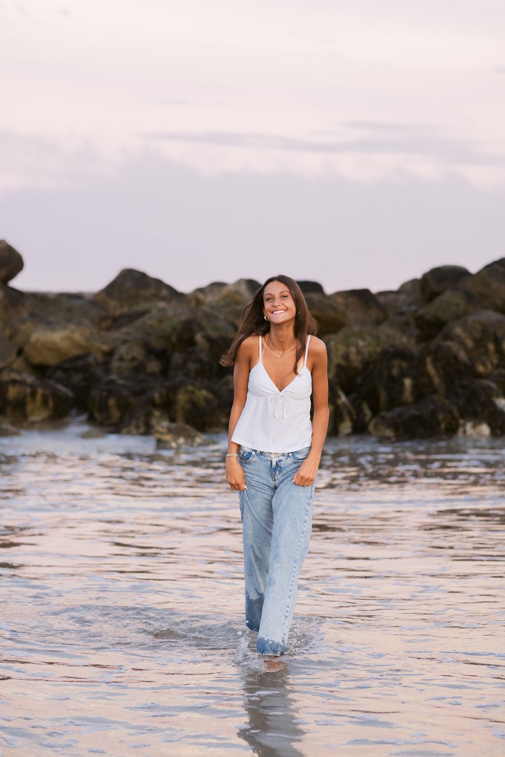 Young lady with a white tank top and blue jeans walks through the ocean at sunset for senior portraits. Kennebunkport senior photographer
