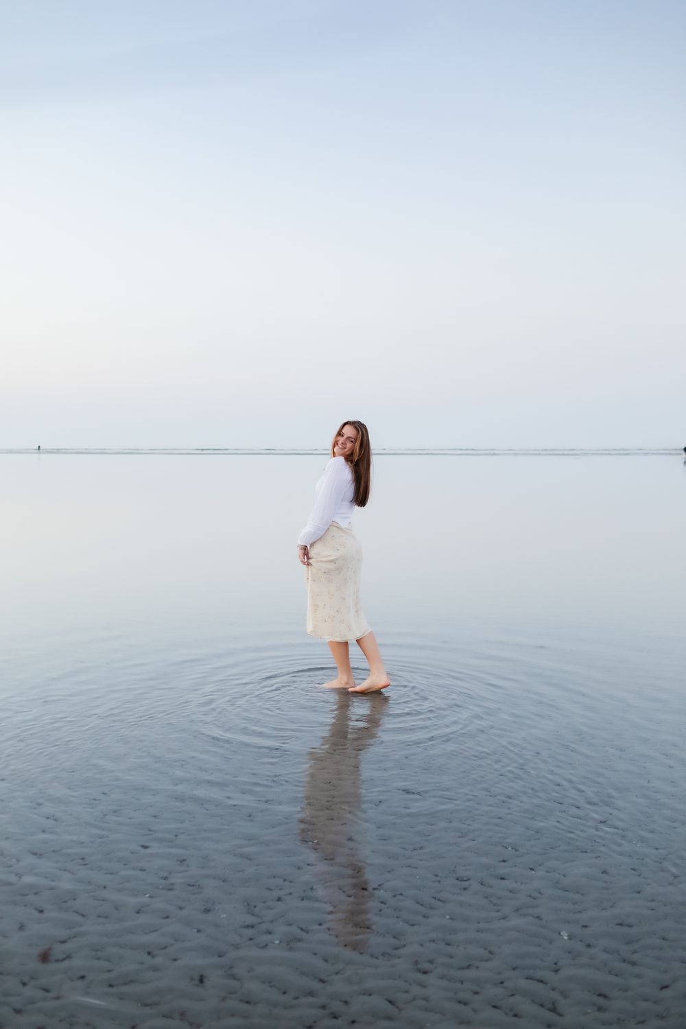 Blond girl plays with her yellow floral skirt in the shallow ocean water of Wingaersheek Beach in Gloucester North Shore for senior photos.