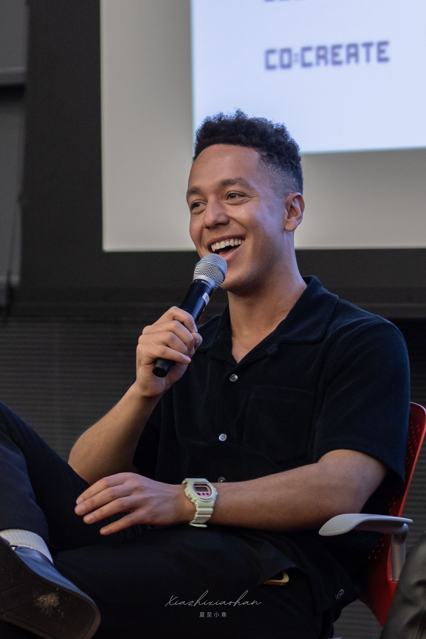 Speaker wearing a black polo shirt smiles while speaking at a conference.