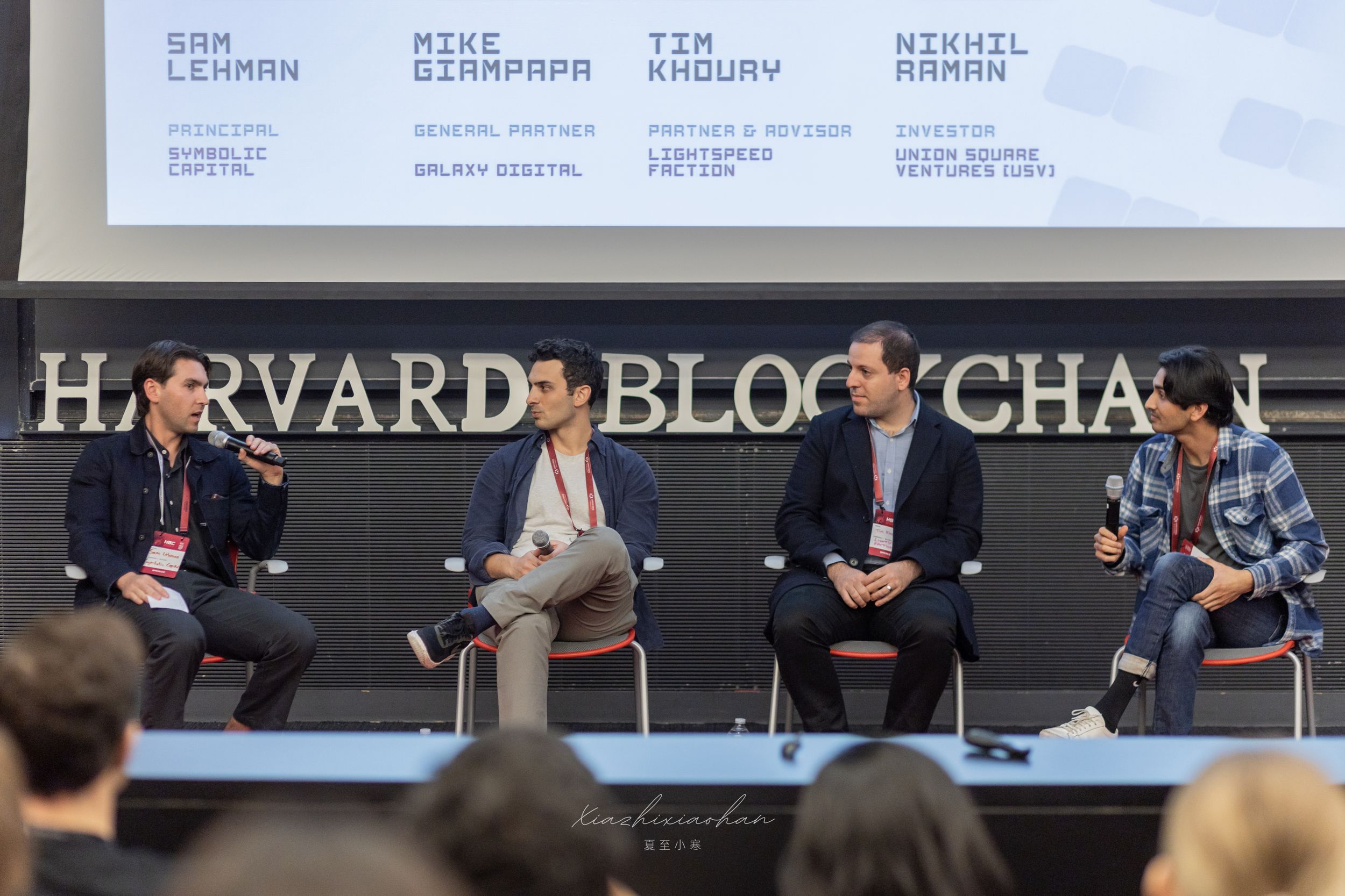 Business conference attendees seated in a row at a Harvard Blockchain panel discussion.
