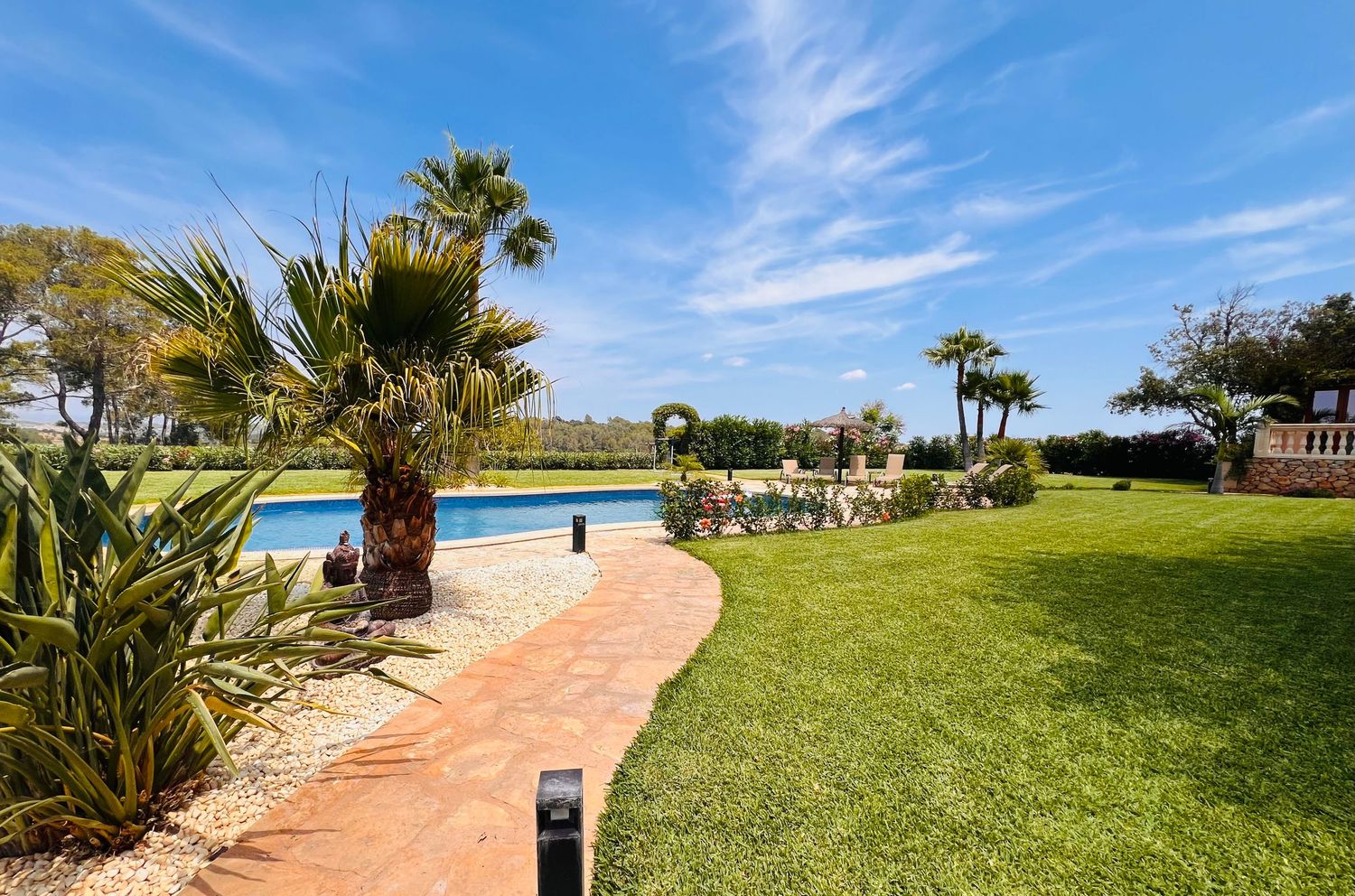 Curved pathway leads to a swimming pool surrounded by palm trees and manicured lawn under bright blue skies.