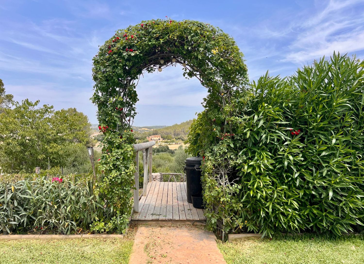 A lush garden archway with climbing vines frames a wooden bridge pathway leading to scenic rolling hills.