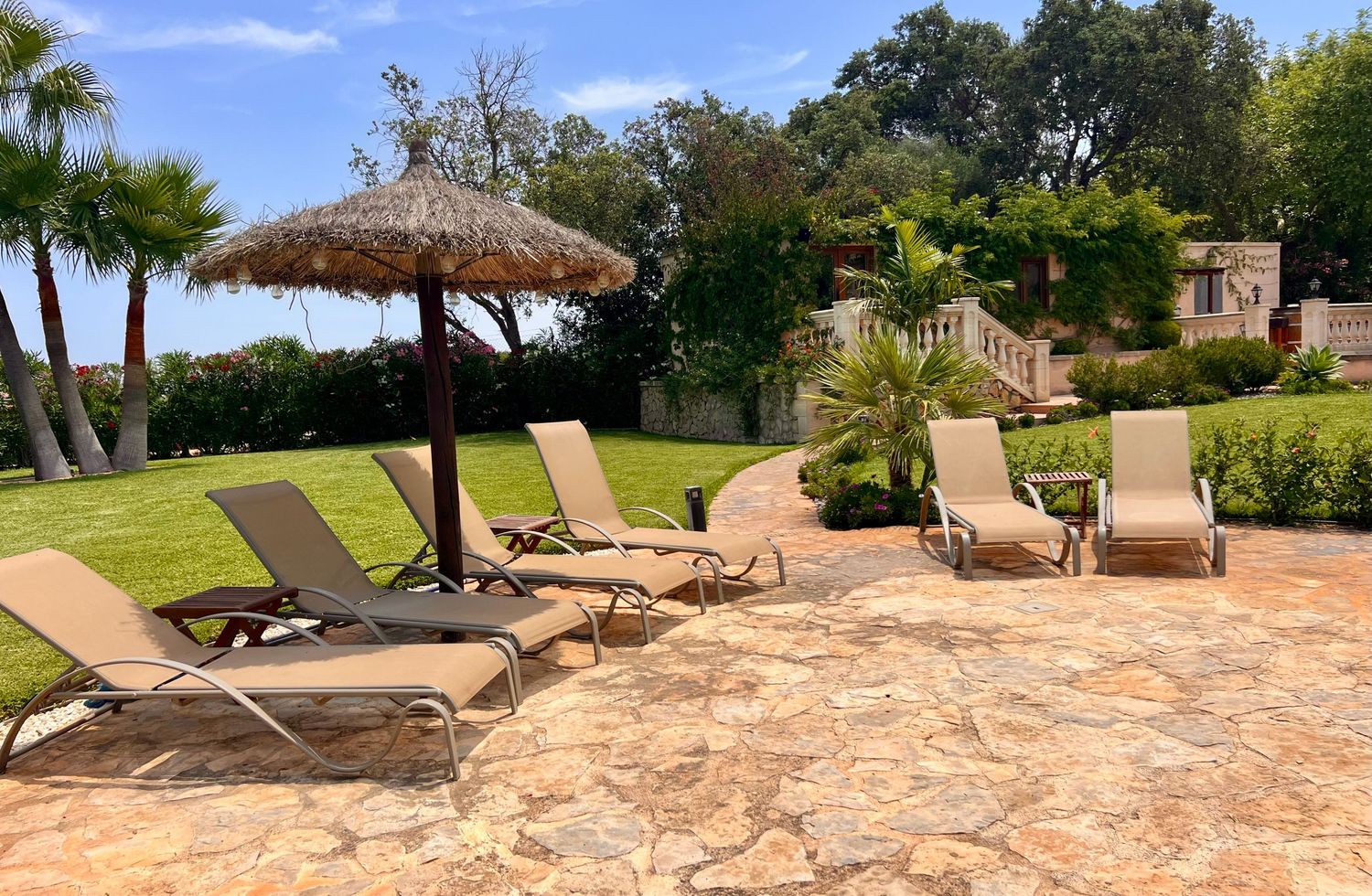 Lounge chairs and tiki umbrella on stone patio overlooking tropical landscaped garden on sunny day.