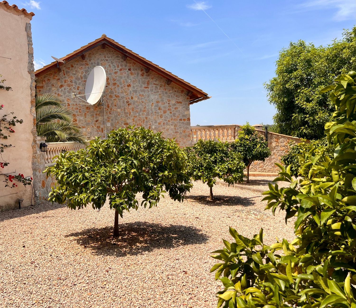 Mediterranean-style stone house with citrus trees in a gravel courtyard under bright blue sky.